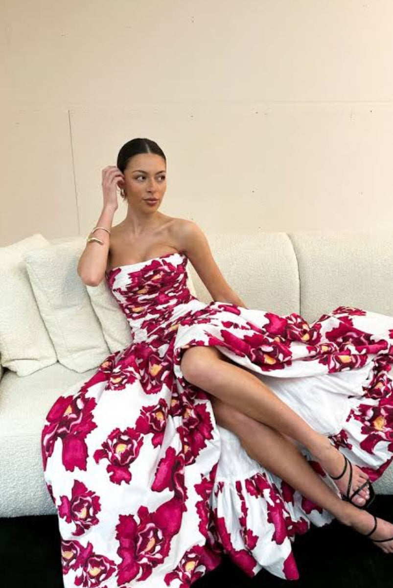 Aje Behold Dress Boldly Blooming Dress in pink and yellow floral on white fabric, model is seated on cream sofa.